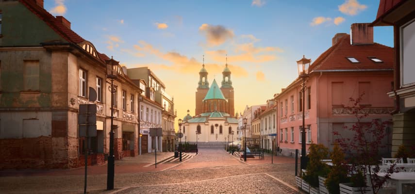 A cobblestone street in Gniezno leading to the twin-towered cathedral at golden hour, framed by pastel townhouses on either side.