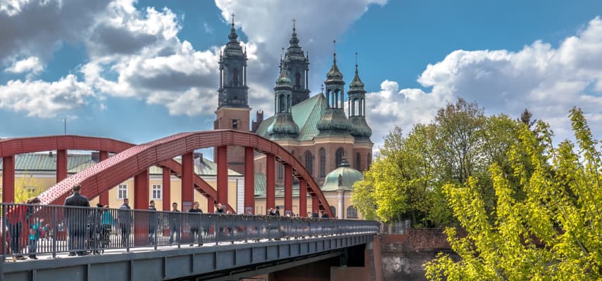 The red arch of Bishop Jordan Bridge leading pedestrians across the Warta toward the four green-domed towers of Poznań Cathedral on Ostrów Tumski.